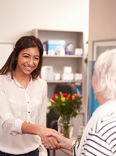 A young woman with dark hair shakes hands with an elderly woman in the lobby of a healthcare office.